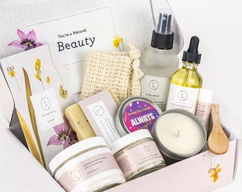 May include: A gift box filled with self-care products, including a lavender body scrub, a clay facial mask, a scented soy candle, a body oil, a mist, and a washcloth. The box is decorated with dried flowers and the text "You're a Natural Beauty".