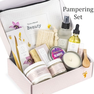 May include: A pink and white gift box filled with a variety of bath and body products, including a candle, body scrub, body oil, and a bath mitt. The box is labeled "Pampering Set" and includes a card that says "You're a Natural Beauty".