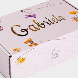 May include: A light purple gift box with a floral design and the name "Gabriela" written in gold lettering. The box has a white label with the letter "L" in a circle and the word "Lizush" underneath.