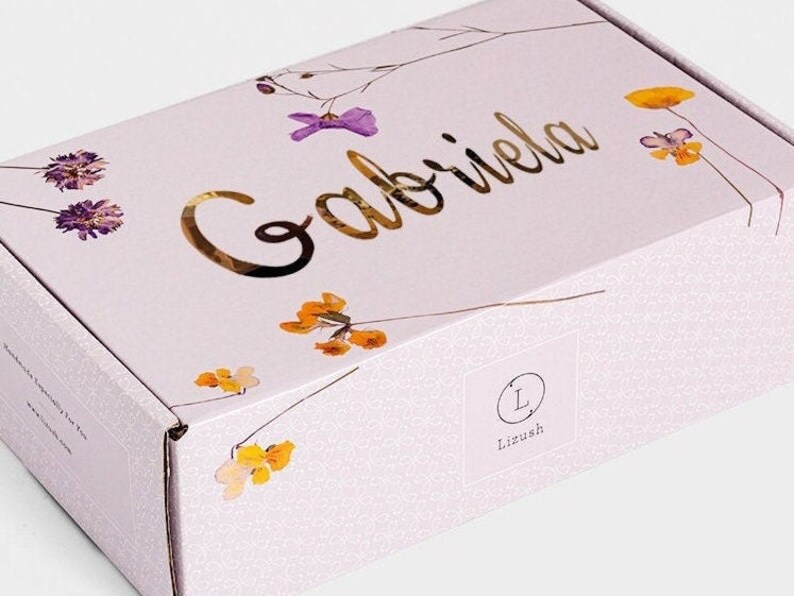 May include: A light purple gift box with a floral design and the name "Gabriela" written in gold lettering. The box has a white label with the letter "L" in a circle and the word "Lizush" underneath.
