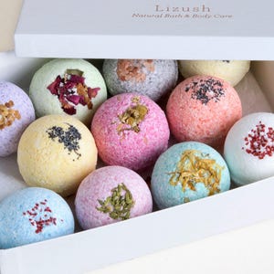 May include: A white box filled with ten colorful bath bombs. The bath bombs are round and have different colors, textures, and ingredients. The box has the text "Lizush Natural Bath & Body Care" printed on the top.