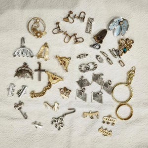 May include: A collection of vintage jewelry pieces, including earrings, charms, and a keychain. The assortment features gold and silver tones, with designs such as flags, crosses, and floral motifs. The items are arranged on a white, textured surface.