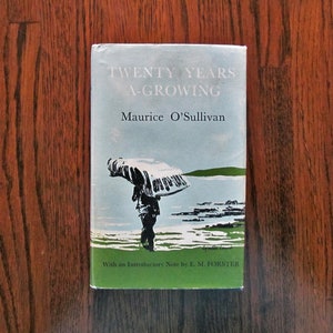 May include: A vintage book titled "Twenty Years A-Growing" by Maurice O'Sullivan. The cover features a person with an umbrella on a beach, with a green border. The book is on a wooden surface.