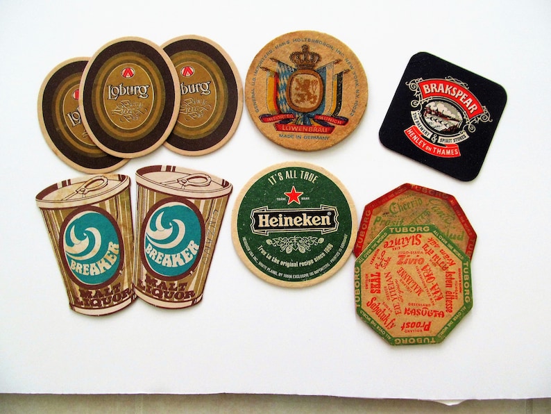 May include: A selection of beer coasters with various brand designs. The coasters are in different shapes, including round and square. Brands include "Loburg", "Heineken", and "BrakSpear". The colours are brown, green, and black.