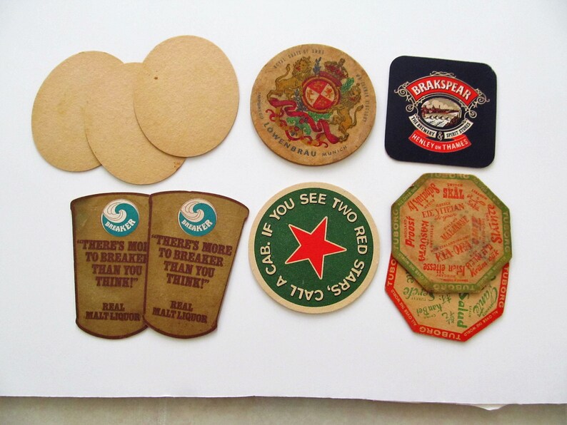 May include: A selection of vintage beer mats in different shapes and designs. Includes round, oval, and square mats with brewery logos and text. Some feature names like Lowenbrau and Brakspaer, and slogans such as "There's More to Breaker Than You Think!"