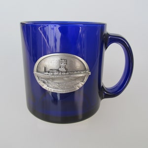 May include: A cobalt blue glass mug with a silver-colored emblem featuring a ship and the text "U.S.S. RONALD REAGAN." The mug has a large handle and a simple, elegant design, suitable for beverages.