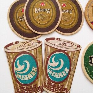 May include: A selection of vintage beer mats and malt liquor cutouts. The mats have a brown and gold design with the word "Loburg". The malt liquor cutouts are shaped like cans with the word "Breaker" and a teal wave design.
