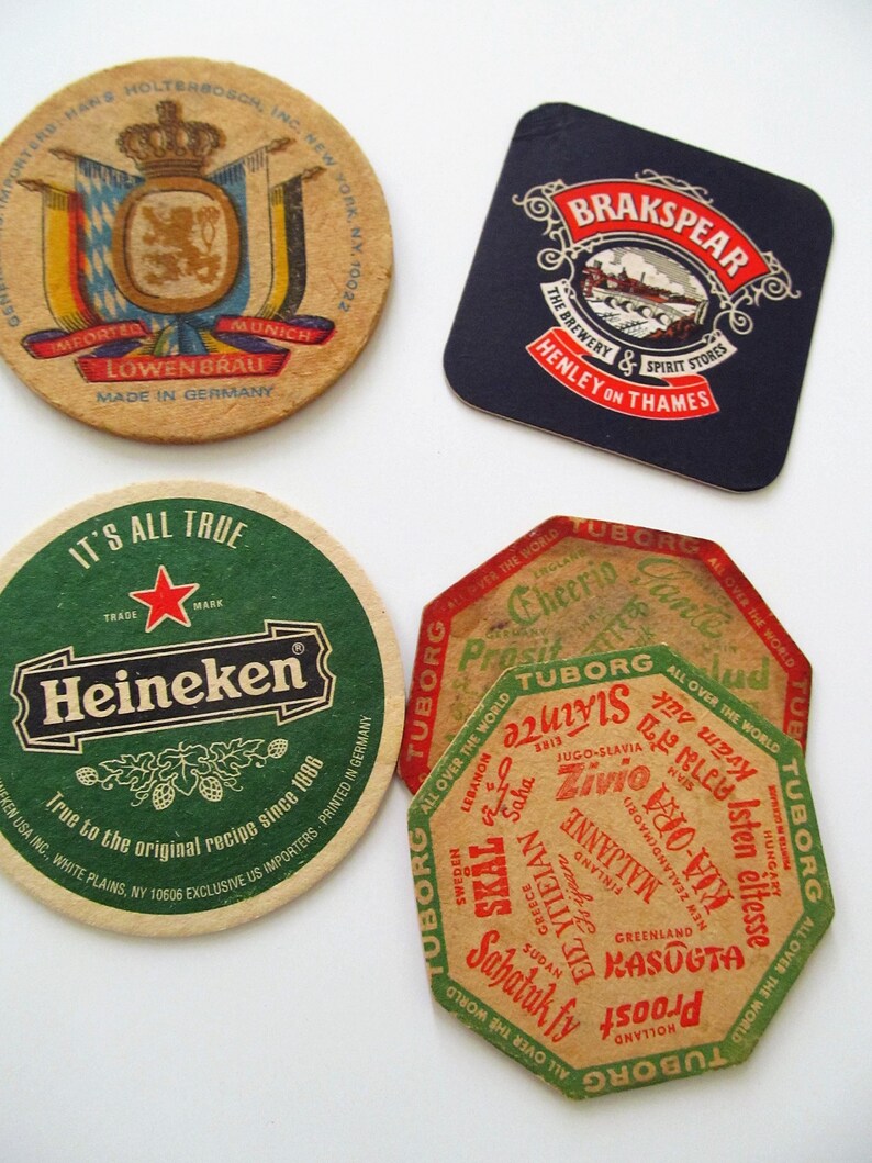 May include: A selection of vintage beer mats with brewery branding. The mats display logos and text for Lowenbrau, Brakspear, Heineken, and Tuborg. The shapes vary, including round, square, and octagonal, with diverse colors and designs.