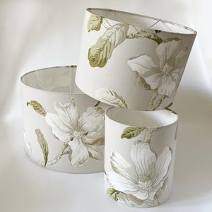 May include: Three lampshades featuring a floral design. The lampshades are made from a beige fabric with white magnolia flowers and green leaves.