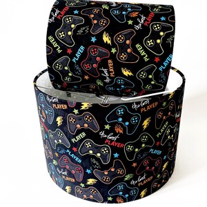 May include: Two black lampshades with a repeating pattern of colourful video game controllers, stars, and lightning bolts. The words "PLAYER" and "the best" are also featured in the design. The lampshades are cylindrical.