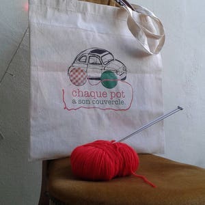 Può includere: Una borsa di tela bianca con un'illustrazione in bianco e nero di un'auto e il testo "chaque pot a son couvercle." Un gomitolo di lana rosso e ferri da maglia sono su una sedia marrone.