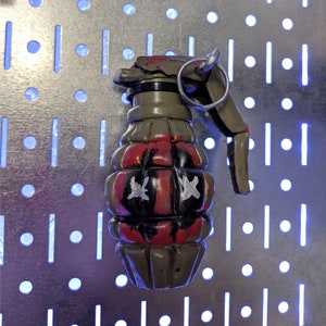 May include: A hand grenade painted in olive green, red, and black. The grenade features white star designs and a silver pull ring. The design has a distressed, worn appearance.