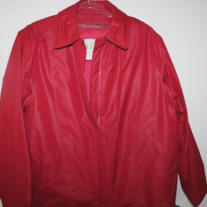May include: A bright red jacket featuring a classic collar, long sleeves, and a central zipper closure. The garment includes a visible interior label near the neckline and is constructed with a smooth, solid-colored fabric throughout the exterior.