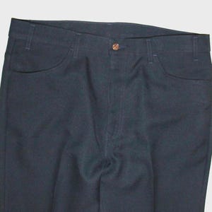 May include: Close-up of dark blue denim pants. The pants have two front pockets and a button closure. The fabric appears to be a medium weight denim. The pants are folded and laid flat.