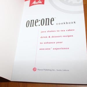 May include: Open cookbook page with the title "one:one cookbook" in black font. The page includes text in red font: "java shakes to tea cakes: drink & dessert recipes to enhance your one:one experience". The Melitta logo is in the upper left corner.