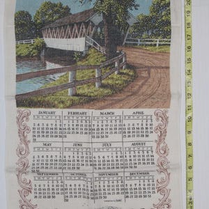 May include: A vintage calendar towel featuring a detailed illustration of a covered bridge over a river, with a dirt road and trees. The calendar displays the year 1982, with months and dates. The towel measures approximately 24 inches in length.