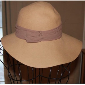 May include: A tan-colored fedora hat with a wide brim and a brown ribbon band. The hat is displayed on a black wire mannequin head. The hat's crown is slightly indented, and the ribbon band is gathered.