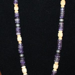 May include: A beaded necklace featuring a repeating pattern of purple amethyst disc beads, cream-colored carved cylindrical beads, and small dark grey metallic spheres. The beads are strung together in a continuous strand on a flexible cord.