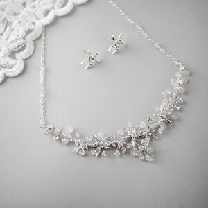 May include: A silver necklace with a delicate chain and a floral design featuring small sparkling crystals. Matching earrings with a star-shaped design are also included.
