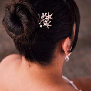 May include: A woman with dark hair styled in a bun with a gold starfish hair comb with pearl accents.
