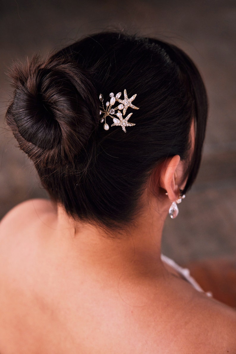 May include: A woman with dark hair styled in a bun with a gold starfish hair comb with pearl accents.