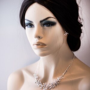 May include: A silver necklace with a delicate floral design featuring small, clear crystals. The necklace is worn by a mannequin with dark hair and makeup.