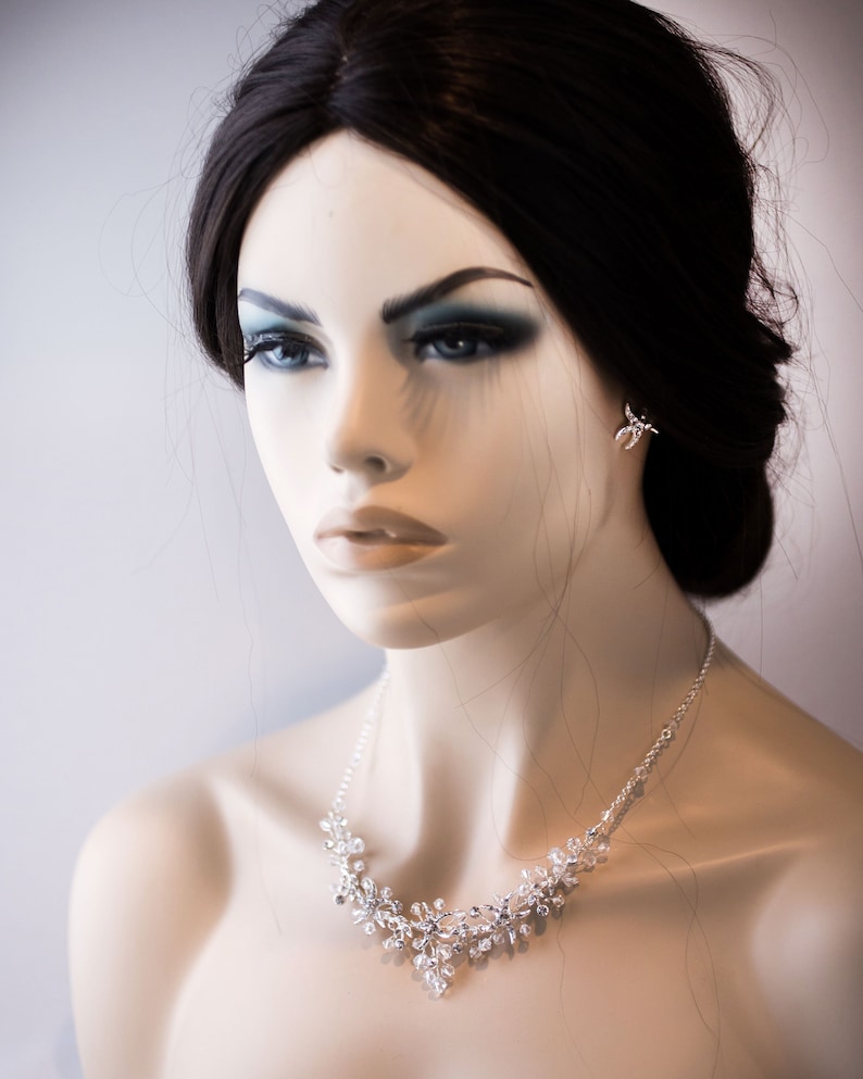 May include: A silver necklace with a delicate floral design featuring small, clear crystals. The necklace is worn by a mannequin with dark hair and makeup.