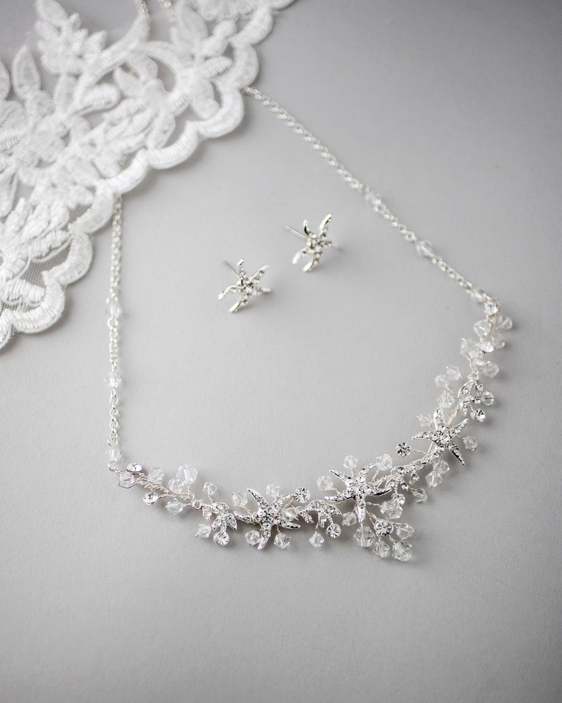 May include: A silver necklace with a delicate chain and a floral design featuring small sparkling crystals. Matching earrings with a star-shaped design are also included.