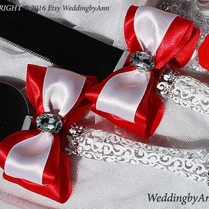 May include: A pair of white and red satin ribbon bows with rhinestone accents, attached to a pair of white lace-wrapped cake cutting knives. The knives are on a white background with the text "WeddingbyAnn" visible.