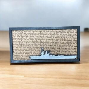 May include: A rectangular wooden plaque with a gray battleship silhouette at the bottom. The background features printed text. The plaque is framed in black and rests on a wooden surface. The overall theme is nautical.