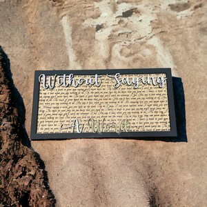 Puede incluir: Un letrero negro enmarcado con las palabras "Without Saying A Word" en una fuente decorativa. El letrero está hecho de madera y tiene un aspecto desgastado. El texto en el letrero es un poema sobre el amor y la conexión.