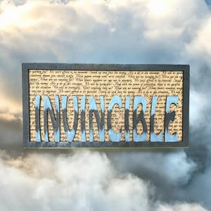Op de afbeelding: Een houten bord met een zwart frame en het woord "INVINCIBLE" in blauw en zwart geschilderd op een achtergrond van tekst. De tekst luidt "We can't afford to be innocent. Stand up and face the enemy. It's a do or die situation. We will be invincible!"