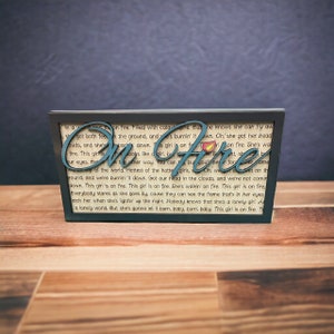 May include: A teal blue wooden sign with the words "On Fire" in large, bold letters. The sign is framed in black and has a white background with text about a girl on fire.