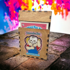 May include: A wooden box with a Garbage Pail Kids card inside. The card features a character with a skull for a head. The box is labeled "Garbage Pail Kids" and "125b" and has a pink background.