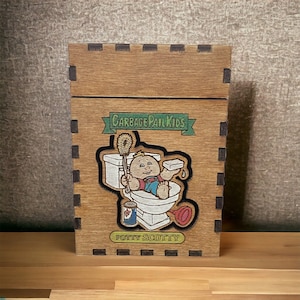 Może przedstawiać: Drewniana skrzynka z kreskówkowym obrazkiem dziecka na toalecie. Skrzynka ma napis "Garbage Pail Kids" i "Potty Scotty".