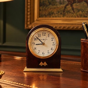 May include: A dark brown, arched wooden mantel clock with a gold-toned bezel and a white face featuring black Arabic numerals. The clock face displays the text Howard Miller. A gold-toned logo is positioned at the base of the clock body.