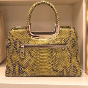 May include: A structured handbag featuring a yellow-green and dark brown snakeskin pattern. The bag has a top zipper closure, a horizontal exterior zippered pocket, and a rounded handle attached with polished silver-tone metal hardware.