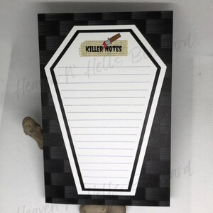 May include: A coffin-shaped notepad with a black and white checkered background. The notepad has a white interior with lined paper and the words "KILLER NOTES" printed on a label with a small knife illustration.