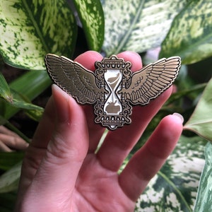 May include: Gold-toned metal pin with an hourglass design and wings. The hourglass is surrounded by an ornate frame.