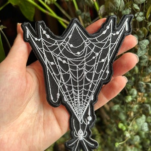 May include: A black and white embroidered patch of a spider web with a spider hanging from the bottom. The web is white and the spider is black.