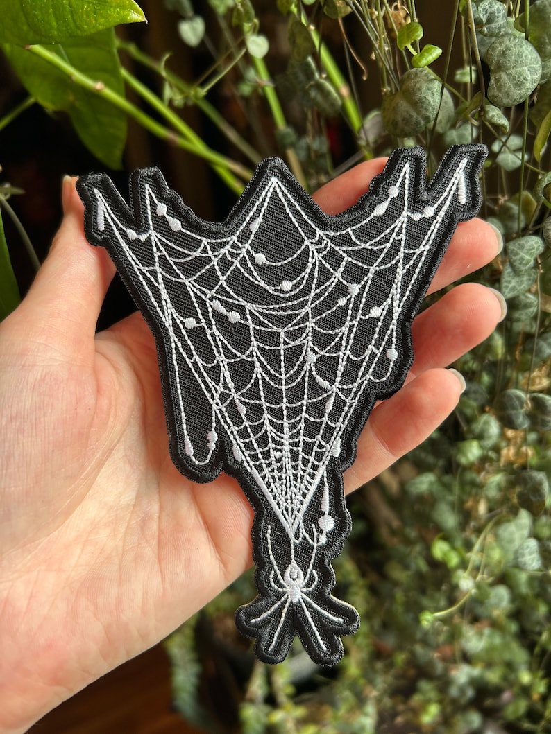 May include: A black and white embroidered patch of a spider web with a spider hanging from the bottom. The web is white and the spider is black.
