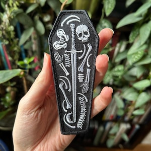 Puede incluir: Parche negro en forma de ataúd con un diseño de esqueleto blanco. El diseño incluye una calavera, huesos y una espada. El parche se sostiene en una mano sobre un fondo de plantas verdes. El parche mide aproximadamente 10 cm de largo.