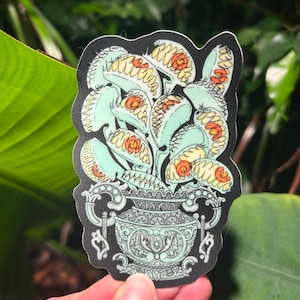May include: A die-cut sticker featuring a bouquet of pale green Venus flytrap plants with orange centers, arranged inside an ornate, light blue decorative urn with intricate scrollwork details and a black background border.
