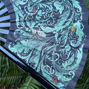 May include: A folding hand fan with a black frame and a dark gray fabric leaf featuring a light green botanical pattern of swirling leaves and vines. The design includes detailed illustrations of various insects, including a beetle and ladybug.