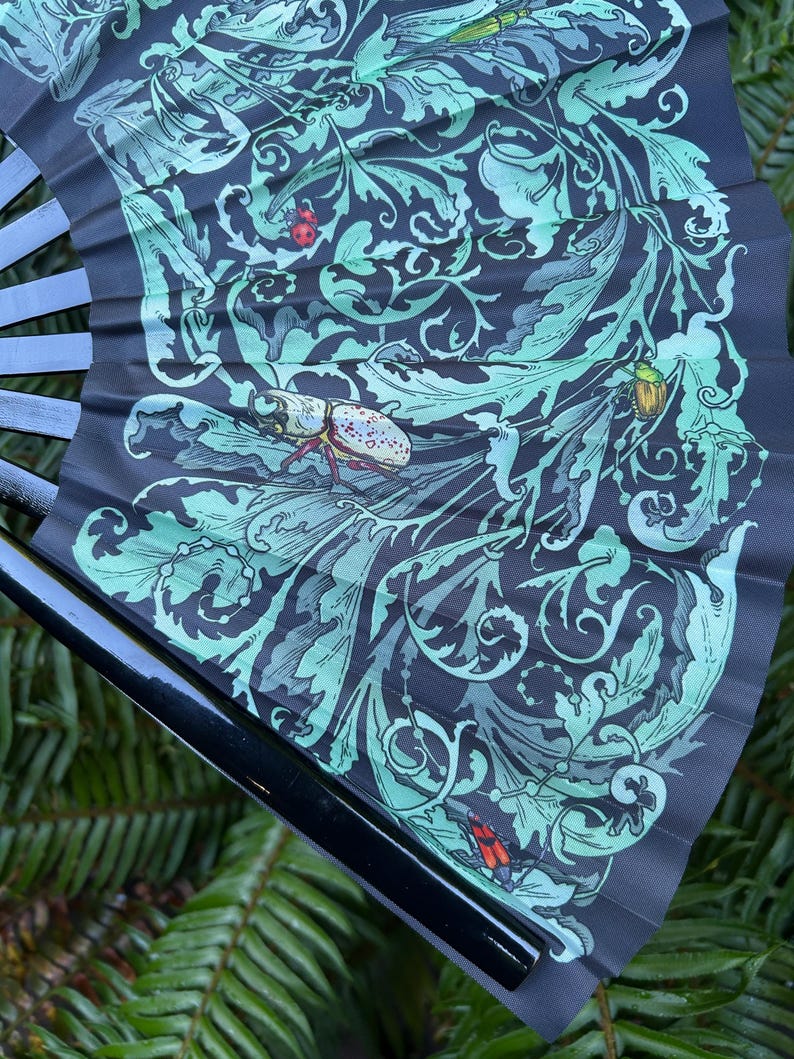 May include: A folding hand fan with a black frame and a dark gray fabric leaf featuring a light green botanical pattern of swirling leaves and vines. The design includes detailed illustrations of various insects, including a beetle and ladybug.