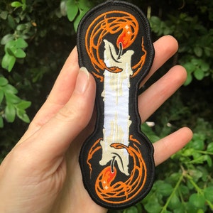 May include: An embroidered iron-on patch featuring a white candle with orange flames and black and orange swirling designs.