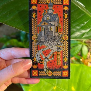May include: A black embroidered patch with a red background and a detailed illustration of a house perched on a clawed, skeletal hand. The house has a pointed roof and windows with glowing lights. The background is decorated with gold and orange floral patterns.