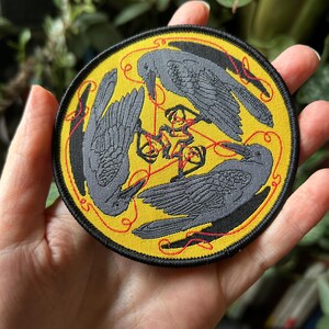 May include: Embroidered patch featuring three gray crows arranged in a circle on a yellow background. Red thread connects the birds and a central design. The patch has a black border.