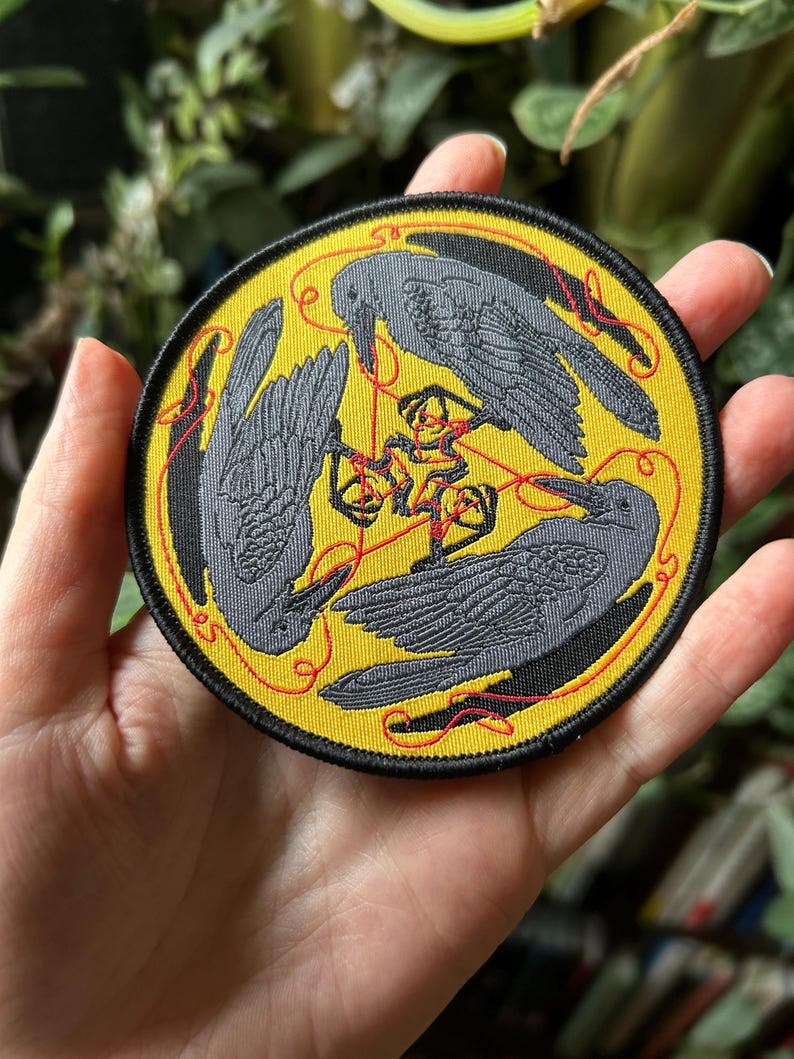 May include: Embroidered patch featuring three gray crows arranged in a circle on a yellow background. Red thread connects the birds and a central design. The patch has a black border.
