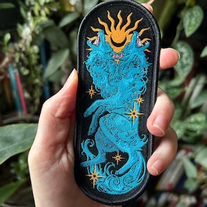 Peut inclure: Écusson brodé ovale noir avec deux loups turquoise hurlant sous un soleil doré. Le motif comprend des étoiles dorées. Le patch a un style artistique détaillé.